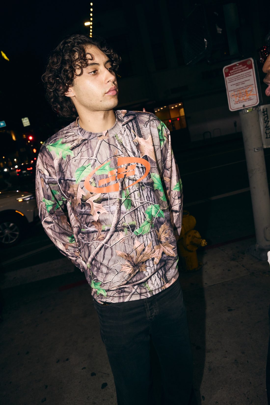 Model in Realtree camo long-sleeve with orange wave graphic, at night on a downtown street
