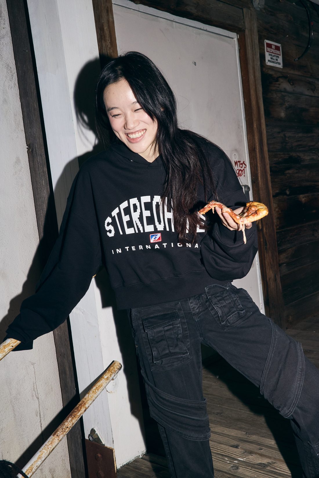 Smiling model leaning at a pizza shop wearing the black STEREOHYPE INTERNATIONAL hoodie